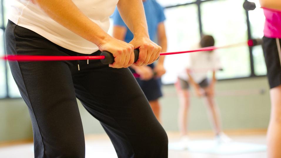 Nahaufnahme einer Person mit einem Widerstandsband im Fitnessstudio, umgeben von weiteren trainierenden Personen.