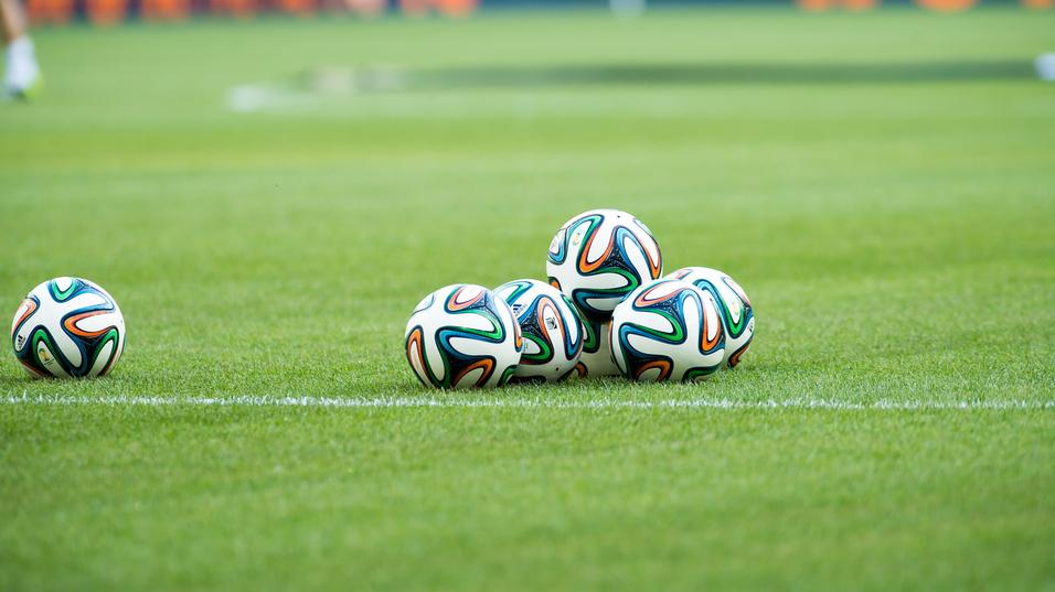 Fünf Fußballbälle liegen auf einem grünen Rasenfeld, sichtbar sind auch der Spielfeldrand und eine verschwommene Hintergrundszene.