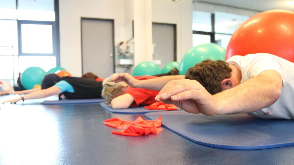 Gruppentraining mit Personen liegend auf Matten, während Bälle im Hintergrund sichtbar sind.